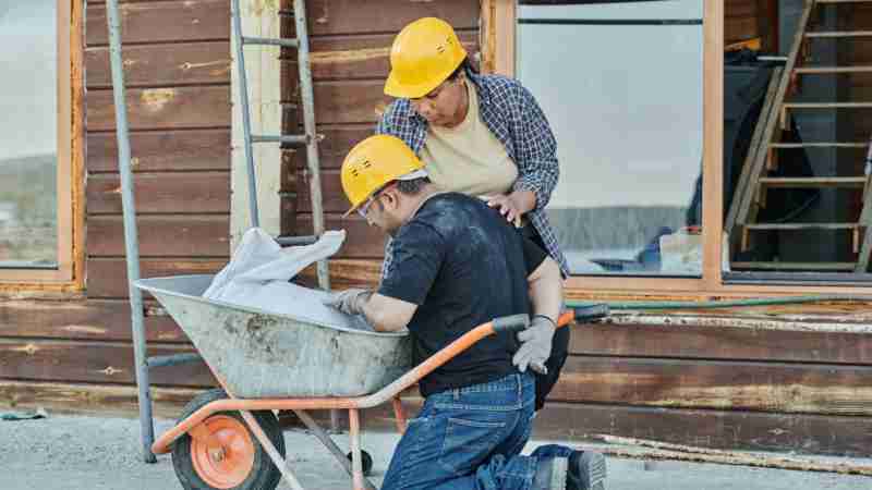 A construction worker receiving help while kneeling with a wheelbarrow on a job site., tags: en - pexel