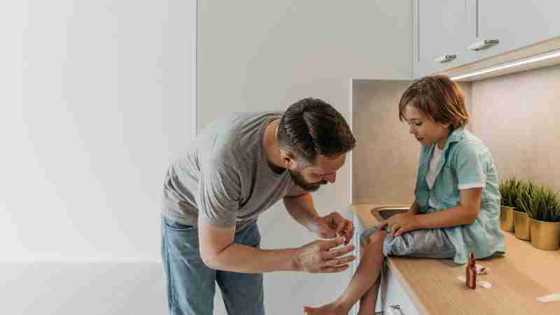 A father is applying a bandage to his son's knee as they sit on a kitchen countertop., tags: kan - pexel
